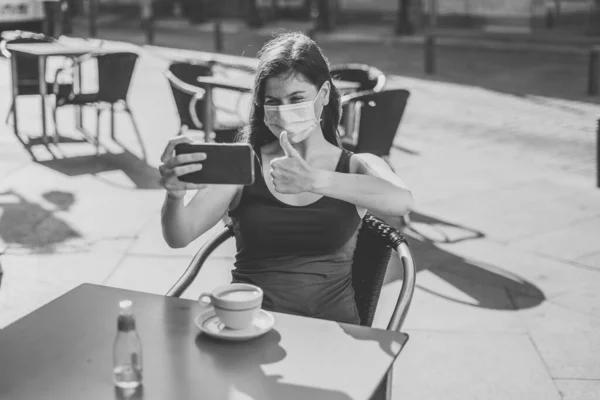 COVID-19 outbreak. Young woman wearing protective surgical face mask and using mobile phone showing thumbs up gesture in a coffee shop terrace. Coronavirus New Normal and mandatory use of face mask