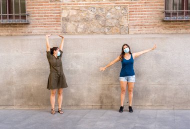 Mutlu kız arkadaşlar koruyucu yüz maskesi takıyor. Birbirlerinden 2 metre uzakta duruyorlar. Sosyal mesafeyi koronavirüsün yayılmasını önlüyorlar. COVID-19 hastalıkların korunması ve Yeni Normal.