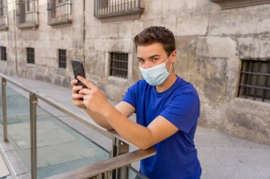 COVID-19 salgını. Şehirdeki genç adam koruyucu cerrahi maske takıyor ve arkadaşlarını ve ailesini cep telefonuyla arıyor. Yeni Normal ve zorunlu yüz maskesi kullanımında bağlı kalın.