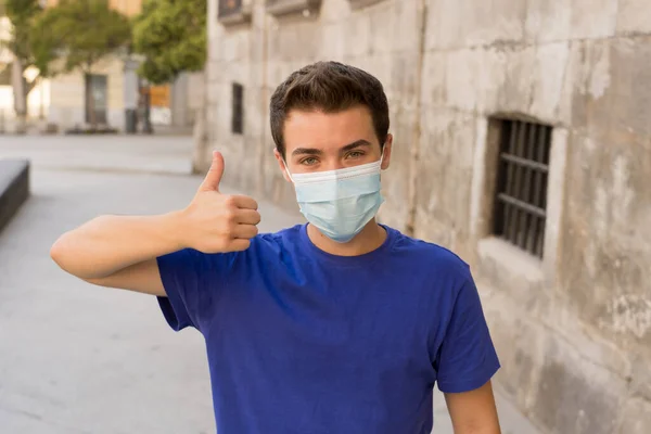 Happy young man with surgical face mask outdoors showing thumbs up. Man with protective mask walking in city street after coronavirus outbreak lockdown. Positive image New Normal life and COVID-19.