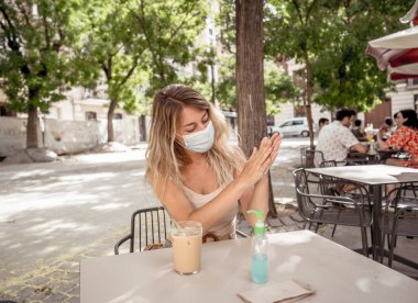 Koruyucu yüz maskesi takan, el dezenfektanı kullanan, New Normal şehir hayatında açık terasta kahve içen genç bir kadın. COVID-19 Salgını, virüsün yayılmasını durdurun, güvende kalın ve sağlık protokolü.