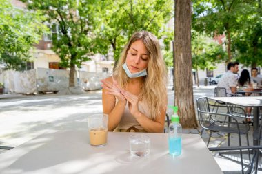 Young woman wearing protective face mask using hand sanitizer drinking coffee in outdoors terrace in the New Normal city life. COVID-19 Outbreak, stop the virus spread, stay safe and health protocol. clipart