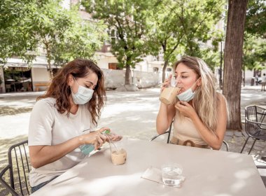 Kahve dükkanındaki kız arkadaşlar koruyucu yüz ve maske takıp el dezenfektanı kullanıyorlar. Coronavirus New Normal 'da içki içen kadınlar. COVID-19 Salgın ve kamu alanlarındaki sağlık protokolleri.