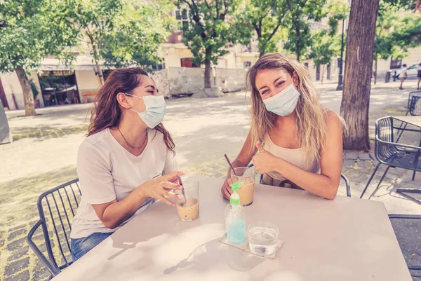 Girlfriends in coffee shop wearing protective face and mask using hand sanitizer. Women having drinks outdoors in the Coronavirus New Normal. COVID-19 Outbreak and health protocols in public spaces.