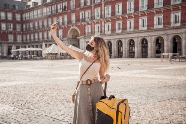 Güzel beyaz kadın koruyucu yüz maskesi takıyor. Plaza Mayor Madrid 'de mutlu ve heyecanlı bir şekilde selfie çekiyor. COVID-19 ve Yeni Normal yaşam tarzı, turizm ve Avrupa seyahatlerinde.