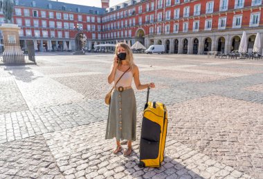 Covid-19 'un turizm üzerindeki etkisi. Madrid 'deki endişeli bir turist, yüz maskesi takıyor ve kişisel izolasyon seyahatleri sırasında cep telefonuyla konuşuyor. Bagajı olan kadın seyahat kurallarından etkilenmiş..