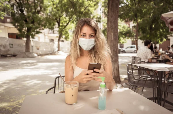 Young woman with protective mask and hand sanitizer on video call using mobile phone in coffee shop in city street. COVID-19 and New Normal, Health safety protocols as mandatory use of face mask.