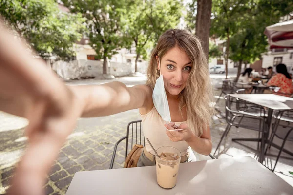 Young woman with protective mask and hand sanitizer on video call using mobile phone in coffee shop in city street. COVID-19 and New Normal, Health safety protocols as mandatory use of face mask.