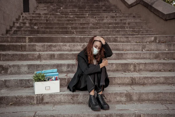 COVID-19 lay off and unemployment. Depressed business woman with face mask outside office with personal staff box feeling hopeless after losing job due to redundancy amid Coronavirus job cuts.