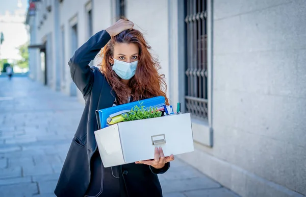 Sad businesswoman in medical protective mask in business district with box of office staff feeling depressed due to job loss. Coronavirus job cuts, COVID-19 unemployment and economic crisis concept.