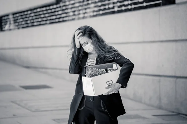 Depressed business woman with face mask outside office with personal staff box feeling hopeless after being fired from work. Coronavirus job losses, lay off and COVID-19 Outbreak impact on employment.