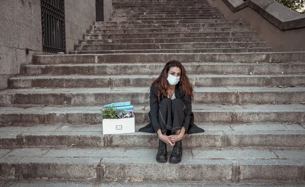 COVID-19 lay off and unemployment. Depressed business woman with face mask outside office with personal staff box feeling hopeless after losing job due to redundancy amid Coronavirus job cuts.