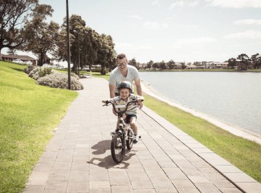 Oğlan babasıyla gölün yanındaki parkta bisiklet sürmeyi öğreniyor. Baba ve oğul bisiklette birlikte eğleniyorlar. Mutlu aile, açık hava aktiviteleri, çocukluk ve ebeveynlik kavramı.