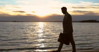 İş adamı el çantasını tutuyor ve gün batımında sahilde yürüyor..