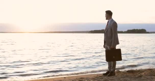 Un homme d'affaires tient une mallette dans sa main et regarde le coucher du soleil — Video