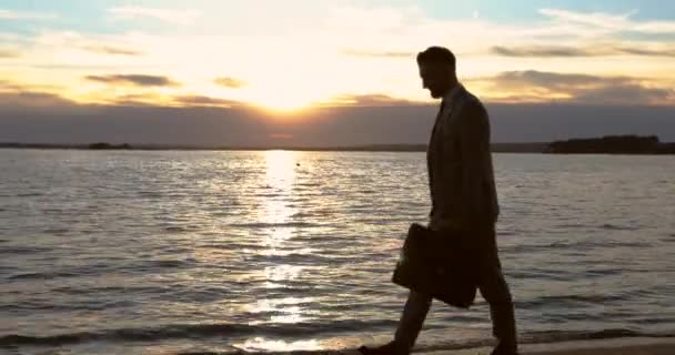 Empresário segurando na mala da mão e andando na praia ao pôr do sol . — Vídeo de Stock