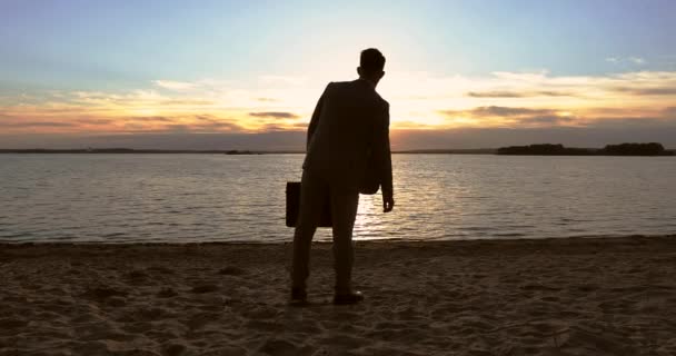 Ένας επιχειρηματίας κρατάει χαρτοφύλακες στα χέρια του και κοιτάζει το ηλιοβασίλεμα δίπλα στη θάλασσα.. — Αρχείο Βίντεο