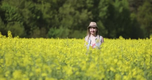 Aantrekkelijke vrouw met een hoed loopt langs de camera in een koolzaadveld in de zomer. — Stockvideo