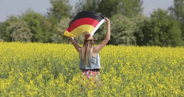 Kobieta z długimi włosami stoi z tyłu i latem trzyma niemiecką flagę na polu rzepaku.. — Wideo stockowe