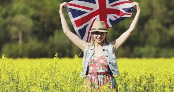 Счастливая привлекательная женщина с английским флагом танцует летом на желтом рапсовом поле. . — стоковое видео