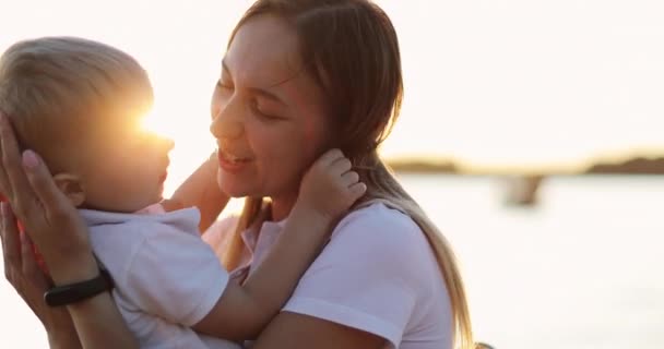 Feliz madre sostiene a un niño pequeño sobre un fondo de una hermosa puesta de sol . — Vídeos de Stock