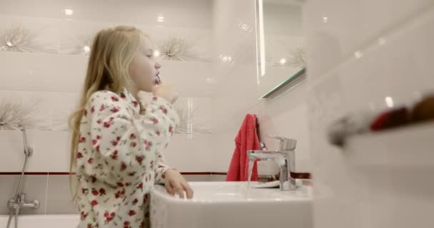 Een schoolmeisje in pyjama poetst haar tanden voor een spiegel. — Stockvideo