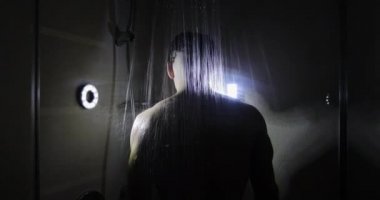 Sakallı bir adam karanlık bir odada duş alıyor..