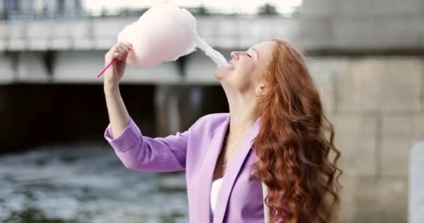 Cheerful girl with bright red hair is eating cotton candy in summer park in time a sunset. — Stockvideo