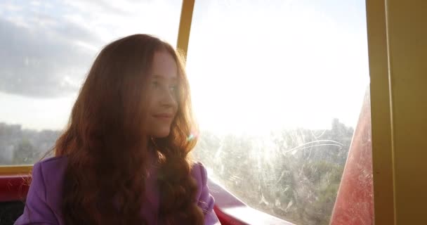 Young woman with curly long red hair is sitting in funicular or ferris wheel cabin in time of sunset. — Stockvideo