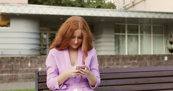 A happy girl with red curly hair is sitting on a bench and texting on mobile phone. — Stockvideo