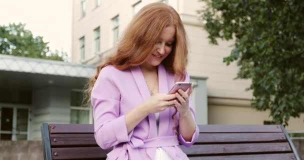 Redhead girl with freckles writes messages on smartphone in the evening outdoors. — Stockvideo