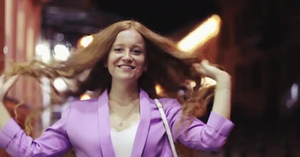 Uma menina feliz com cabelo encaracolado vermelho está andando na cidade da noite . — Vídeo de Stock
