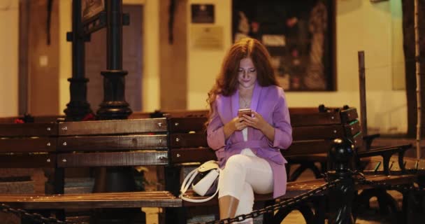 Young woman with red curly hair is typing text message on mobile phone in the night city. — Stock Video
