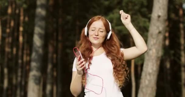 Redhead girl with white headphones is listening songs on mobile phone and dancing with closed eyes. — Stock Video