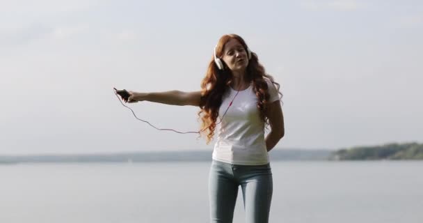 Pelirroja con auriculares blancos está escuchando canciones en el teléfono móvil y bailando . — Vídeo de stock