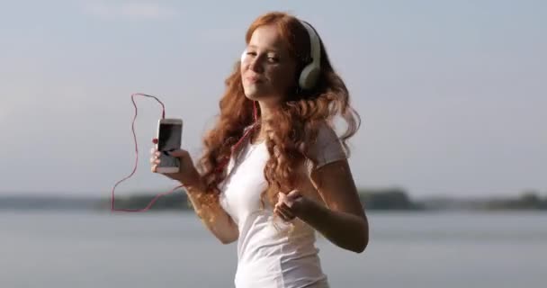 Menina ruiva com fones de ouvido brancos está andando e dançando em uma margem de um rio ouvindo músicas no telefone celular . — Vídeo de Stock