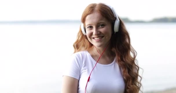 Ansiktet nära. Redhead fräknig flicka med vita hörlurar dansar på en strand av en sjö lyssna låtar. — Stockvideo