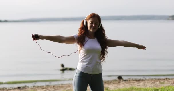 Joven mujer feliz con el pelo rizado rojo disfrutando de la música en los auriculares en una orilla del lago . — Vídeo de stock