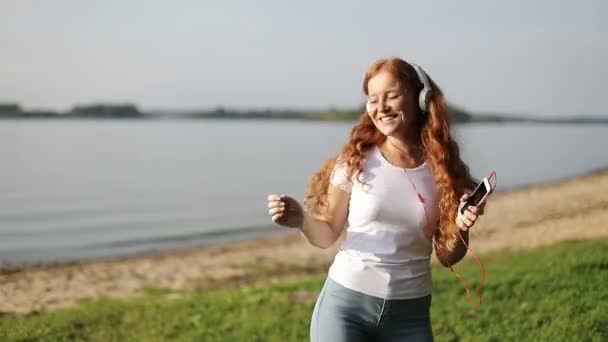 Slow motion. Young happy woman with red curly hair enjoying music and dancing in earphones on a shore of lake. — ストック動画