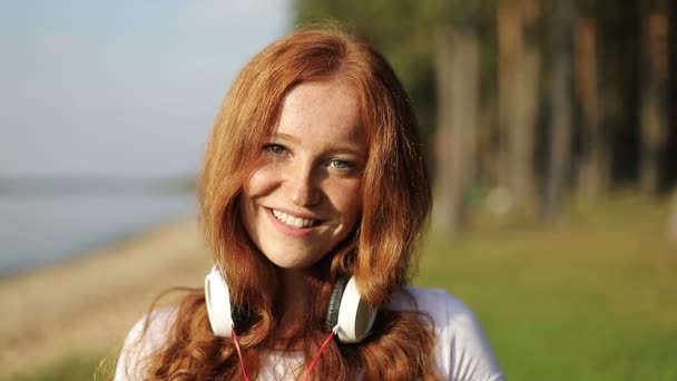 Portrait de jeune femme heureuse avec des cheveux bouclés rouges avec des écouteurs sur le cou . — Video