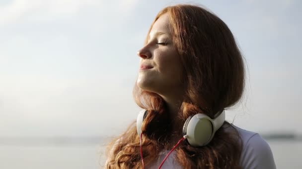 En cámara lenta. Retrato de una joven bailarina feliz con el pelo rizado rojo con auriculares en el cuello . — Vídeos de Stock