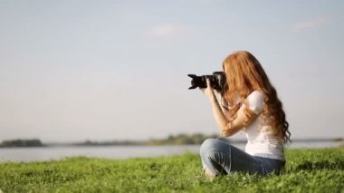 Çilli, çilli, fotoğraf makinesi olan bir kız yeşil çimlerin üzerinde oturuyor ve gölün yakınındaki ormanda fotoğraf çekiyor..