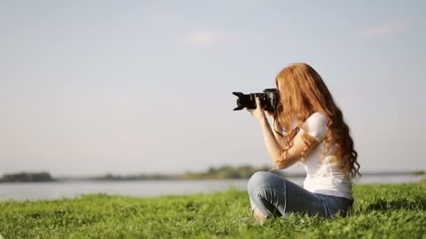 Sun-kissed freckled redhead girl with photo-camera is sitting on a green grass and takes a photo in forest near the lake. — Stock Video