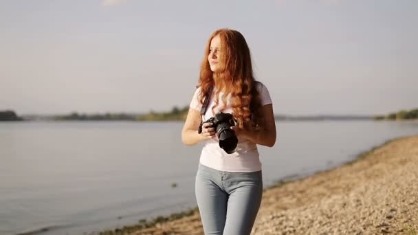 Sun-kissed freckled redhead girl with photo-camera is walking on bank of river. — Stock Video