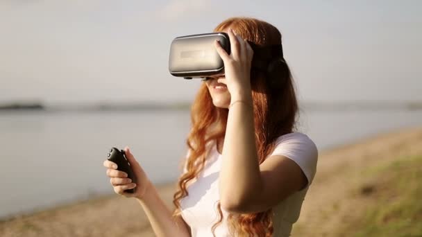 Freckled redhead girl in VR glasses with earphone is watching 360 degrees video using remote control. — Stock Video