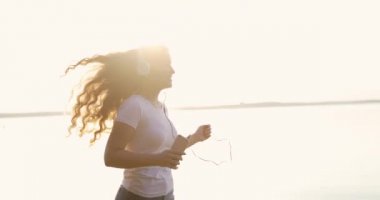 Beyaz kulaklıklı kızıl saçlı kız bir nehir kıyısında koşuyor ve cep telefonundan müzik dinliyor..