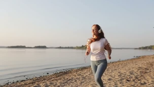 Beyaz kulaklıklı kızıl saçlı kız gölün kıyısında koşuyor ve cep telefonundan müzik dinliyor.. — Stok video