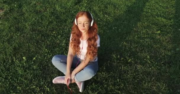Руда веснянка дівчина з білими навушниками слухає пісні і посміхається сидячи на зеленій траві на літньому полі . — стокове відео