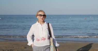 Kulaklıklı, derin kırışıklıkları olan yaşlı bir kadın deniz kıyısında yürüyor.. 