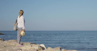 Yaşlı bir kadın sırtı küçük bir kayanın üzerinde duruyor ve denize bakıyor..